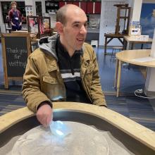 An adult resident sits at a sand table inside the Discovery Museum while holding a small shovel.