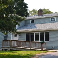 A light green house seen from the driveway with a wooden ramp leading up to the front door.