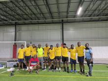 A group of men stand together in front of a goal on an indoor soccer field with their arms around each other wearing matching yellow soccer jerseys.