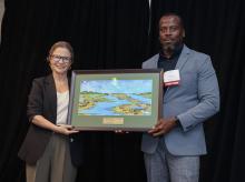 Amy Sousa and Junior Paris stand together in front of a black backdrop, each holding one side of a framed painting of water and marshland.
